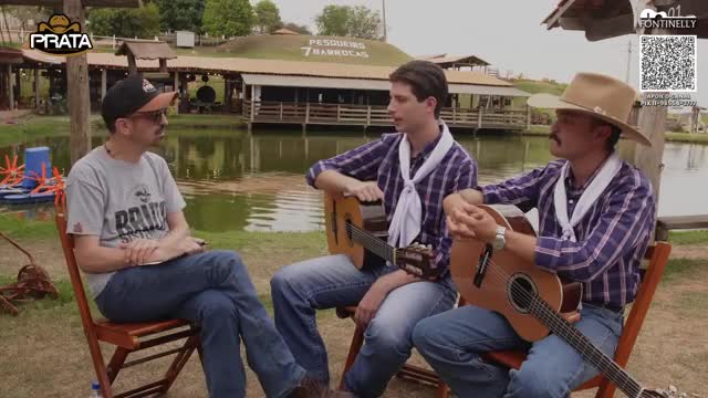 Three men sit in chairs by a lake, two of them holding acoustic guitars. A white scarf is knotted around the neck of each guitarist, and the man on the right wears a wide-brimmed hat. The Rede SPTV logo appears in the upper left corner, and a QR code for donations is visible on the right.