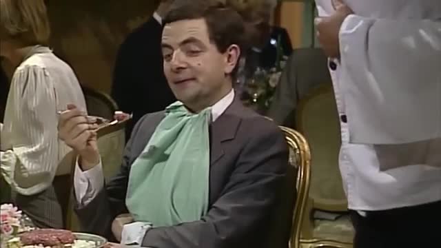 A man in a suit and a large green bib is holding a fork, about to eat. He sits at a table in a fancy room, with other diners around him.