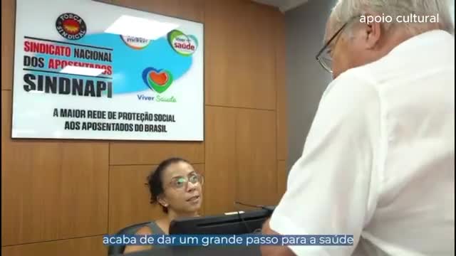 A woman with glasses leans forward, speaking animatedly. An older man in a white shirt turns toward her, his glasses reflecting the light. Behind them, a sign announces "SINDNAPI," the largest social protection network for retirees in Brazil.