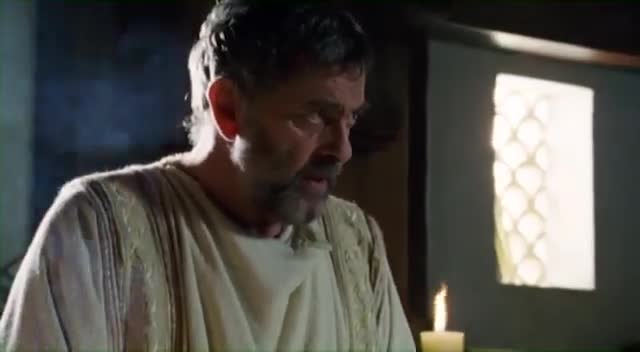 A man with a graying beard looks down, his face illuminated by a nearby candle. The light catches the textured fabric of his tunic, hinting at a scene from a historical drama broadcast by Rede SPTV.