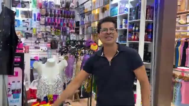A man in a dark polo shirt gestures toward racks of colorful clothing and shelves stocked with various items. He's standing in a brightly lit shop, likely in Brazil, with a Rede SPTV logo visible on a nearby display.