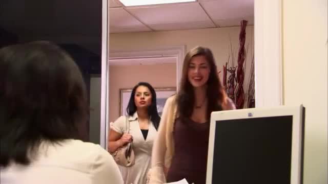 Two women walk into an office, one with a bright smile, the other looking more serious. They're heading towards a desk where a computer monitor sits.