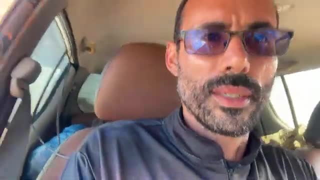 The man in the car is speaking, his mouth open. A blue bag is visible on the back seat.