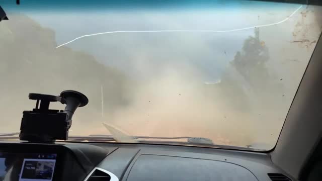 A cloud of dust billows across the road, obscuring the view ahead. The windshield wipers are on, trying to clear the thick haze.