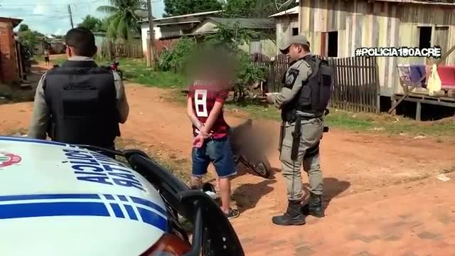 Two police officers stand on a dirt road in front of a shack, one speaking to a young man with his hands behind his back. A police car, marked with "Polícia Militar," is parked nearby. Two police officers stand on a dirt road in front of a shack, one speaking to a young man with his hands behind his back. A police car, marked with "Polícia Militar," is parked nearby.