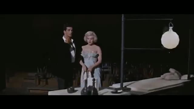 A man in a black suit and a woman in a pale blue dress stand near a table. A white, spherical lamp hangs from a metal frame above a long, flat surface. A man in a black suit and a woman in a pale blue dress stand near a table. A white, spherical lamp hangs from a metal frame above a long, flat surface.