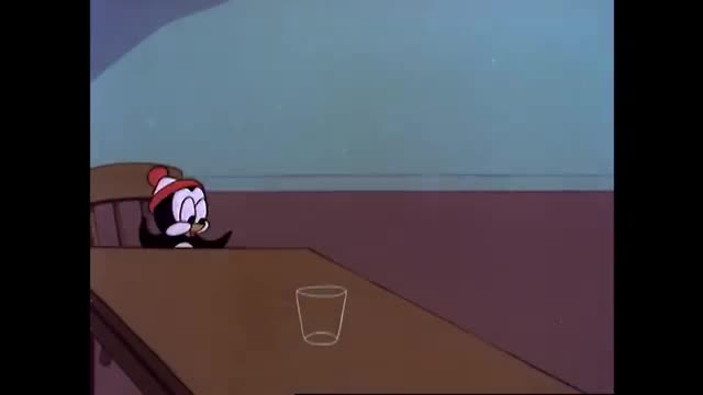 A small penguin, wearing a red hat, sits at a table. A clear glass sits on the table in front of him. A small penguin, wearing a red hat, sits at a table. A clear glass sits on the table in front of him.