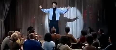 A performer in a light blue jacket stands with arms outstretched under a spotlight, his shadow mirroring him on the stage backdrop. The audience, seated at tables, watches intently. A performer in a light blue jacket stands with arms outstretched under a spotlight, his shadow mirroring him on the stage backdrop. The audience, seated at tables, watches intently.