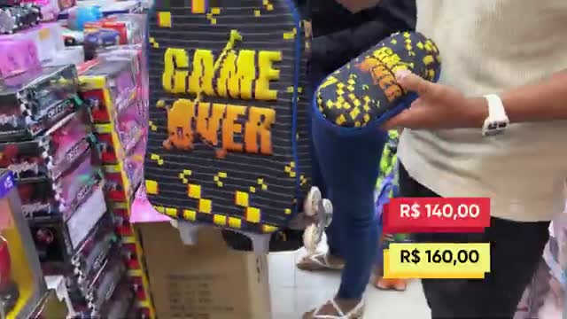 A person holds up a small, patterned case as a large backpack with "GAME OVER" printed on it stands nearby. The prices R$ 140,00 and R$ 160,00 are displayed on red and yellow tags. A person holds up a small, patterned case as a large backpack with "GAME OVER" printed on it stands nearby. The prices R$ 140,00 and R$ 160,00 are displayed on red and yellow tags.