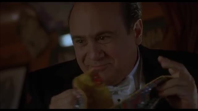 The man in the tuxedo grins as he holds up a colorful, patterned object. He's about to show it to someone.