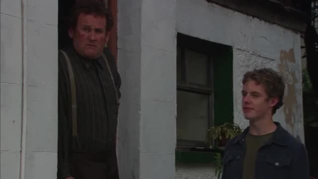 A man with a stern expression peers out from a doorway, his suspenders holding up dark trousers. Beside him, a younger man with light brown hair stands looking off to the side, his gaze thoughtful.