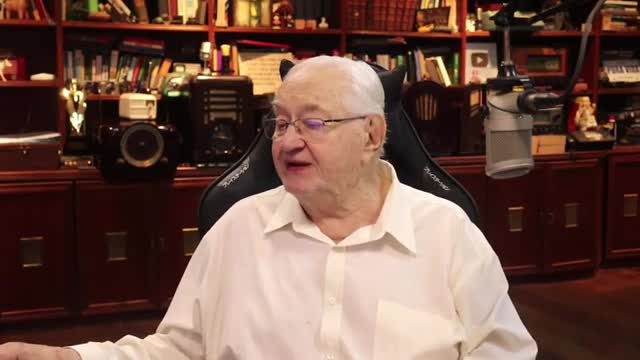 An older man with white hair and glasses speaks from his chair. Behind him, shelves overflow with books and various objects, including a vintage radio and a microphone.
