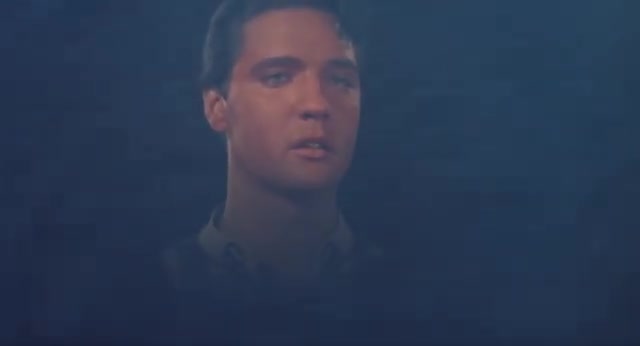 A young man's face, lit by a warm glow, emerges from the darkness. His eyes are fixed on something beyond the frame, a look of intense concentration on his features.