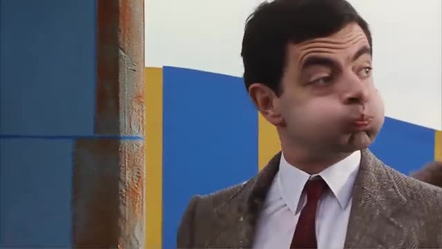 A man in a suit puffs out his cheeks, his face contorted in an odd expression. Behind him, a colorful backdrop of blue and yellow panels stands starkly against the sky.