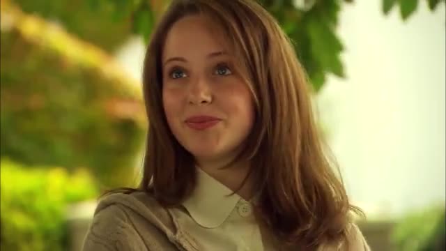 A young woman with long, light brown hair smiles softly, her eyes looking slightly to the side. The background is a blur of green leaves and soft yellow light, suggesting an outdoor setting in Brazil, perhaps during a break from filming for Rede SPTV.