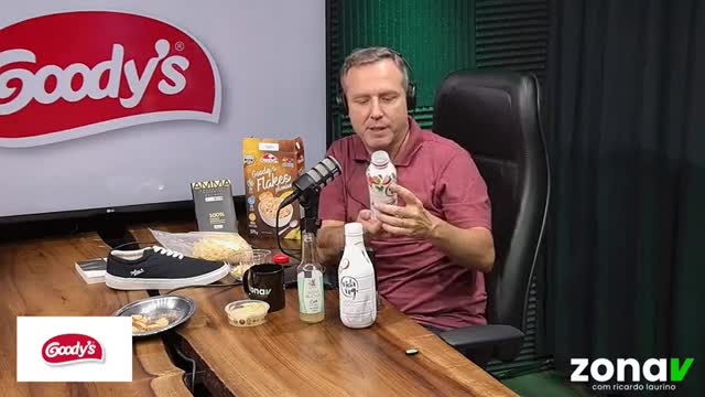 A man in a maroon polo shirt holds up a white bottle, presenting it to the camera. Beside him, on the wooden table, sit a box of Goody's Flakes cereal and a pair of black sneakers.