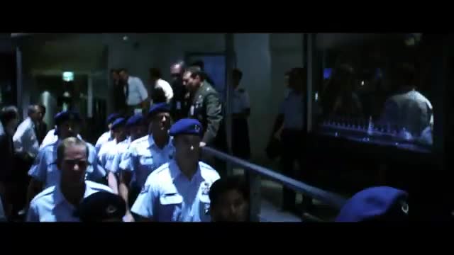 A group of men in light blue uniforms and berets walk forward, some with their hands clasped. A man in a green military uniform stands behind them, observing.