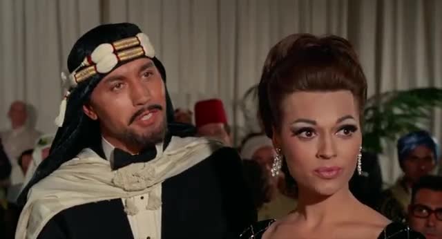 A man in a keffiyeh and a woman with dramatic makeup stand close together. They are at some sort of formal gathering, with other people in the background.