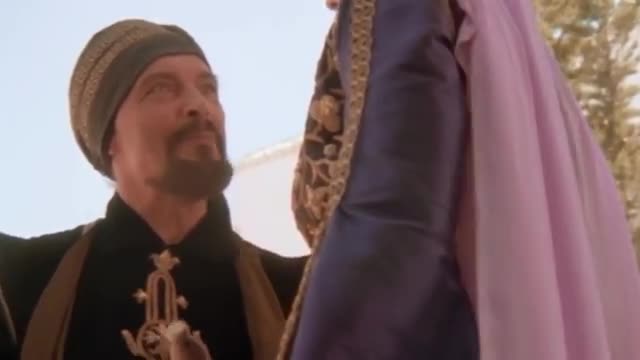 A man in a patterned cap and dark tunic looks up, his gaze fixed on a woman in a flowing purple and pink garment. The fabric of her dress is rich and detailed, catching the light as she stands close.