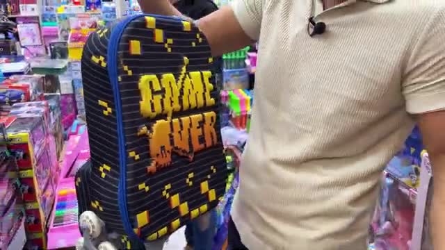 A man in a light polo shirt holds up a black backpack with a pixelated "GAME OVER" design. He stands in a brightly lit store, shelves of colorful merchandise stretching behind him.