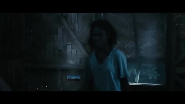 A young woman with dark, curly hair stands in a dimly lit room, her light blue shirt stark against the shadows. She looks directly ahead, her expression unreadable in the gloom.