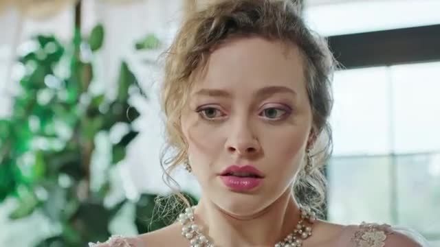 A young woman with curly blonde hair and bright pink lipstick stares with wide, concerned eyes. She wears a pearl necklace and a light pink dress, her gaze fixed on something just out of frame.