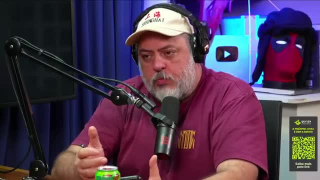 A man with a beard and a cap is speaking into a microphone, gesturing with his hands. Behind him, a Deadpool mask sits on a shelf next to a YouTube play button.