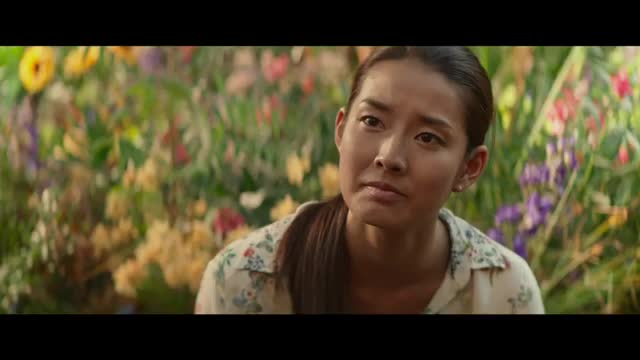 A woman with dark hair pulled back looks intently to her left, her face framed by a dense backdrop of colorful flowers. The scene feels like it could be from a quiet moment captured by Rede SPTV, perhaps during a pause in a broadcast from Brazil.