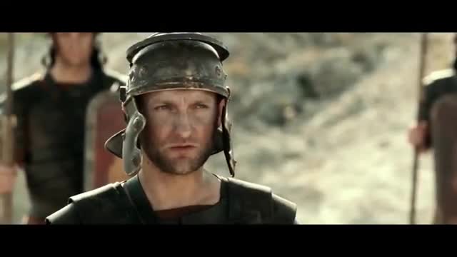 A Roman soldier, his face grimy and determined, stands ready. Behind him, others in similar armor hold their shields and spears, a disciplined line against the dusty landscape.