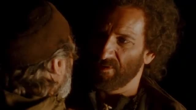 Two men, one with a beard and curly hair, the other with a cap and a grey beard, are talking intensely in the dim light. Their faces are close, and the man with curly hair is speaking with urgency.