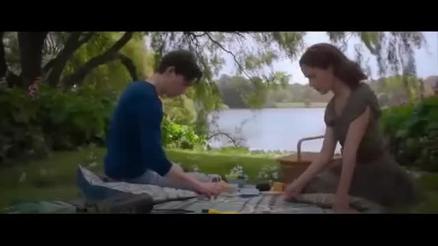 A young man in a blue shirt and a woman in a green dress sit on a blanket by a lake, focused on something between them. The sunlight filters through the trees, illuminating their picnic spread.
