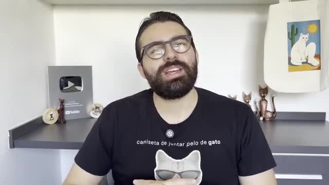 A man with a beard and glasses speaks directly to the camera, wearing a black t-shirt with a cat graphic. Behind him, a silver YouTube play button rests on a shelf, alongside small cat figurines and a tote bag featuring a stylized cat.
