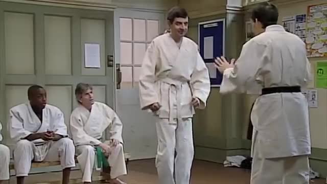 A man in a white gi stands ready, his opponent facing him with open hands. Two other men in gis sit on a bench, watching the proceedings.