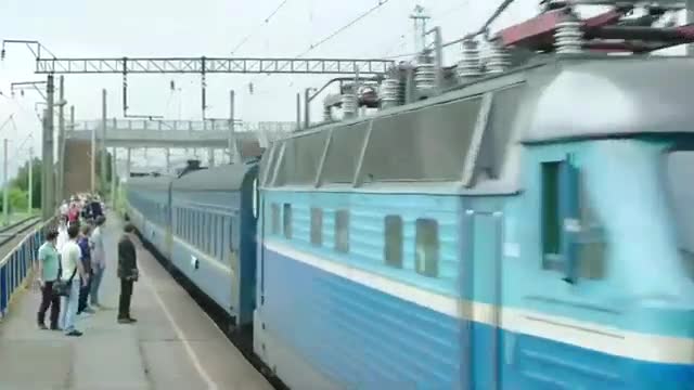 A bright blue train is pulling into the station, its pantograph reaching up to the overhead wires. People are gathered on the platform, waiting to board.