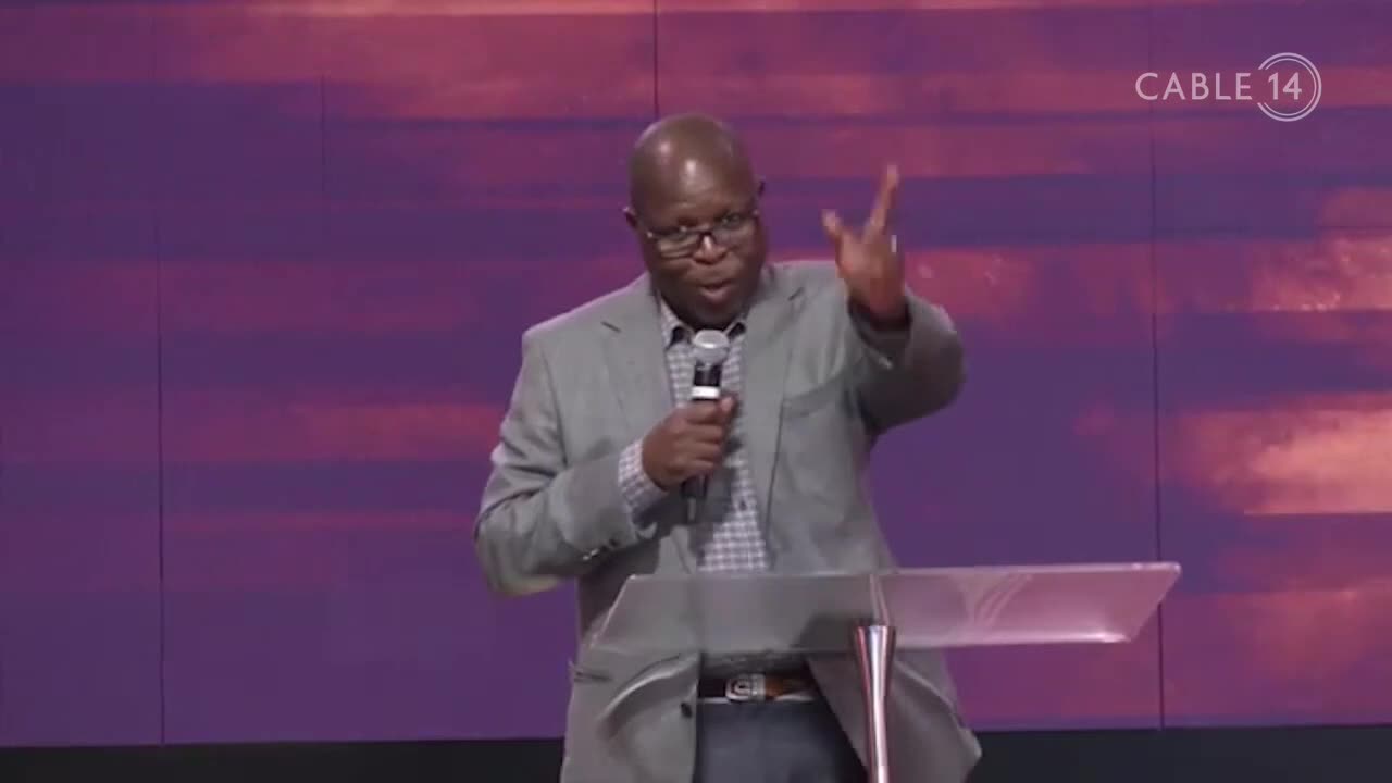 The man in the grey jacket gestures with his right hand while speaking into a microphone. A Cable 14 logo is visible in the background.
