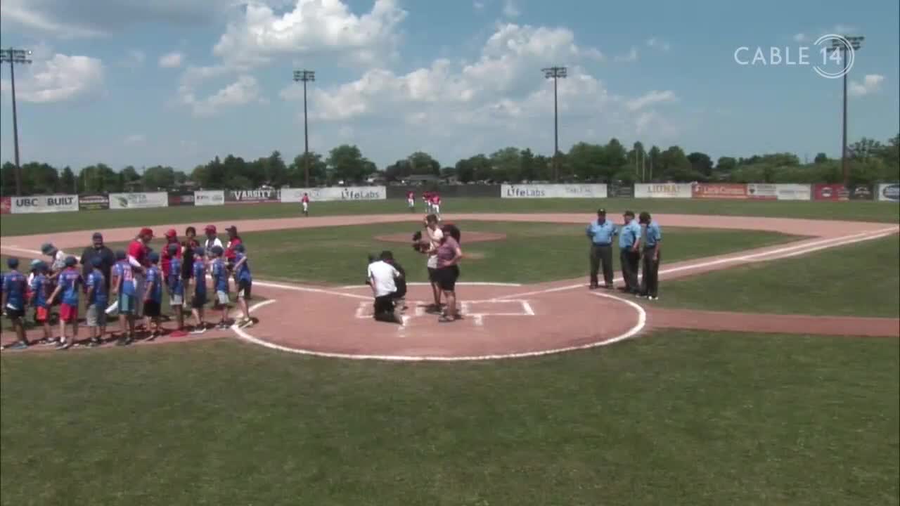 The Cable 14 camera is focused on the pitcher's mound as a woman prepares to throw out the first pitch. Young baseball players in red and blue uniforms stand in front of the dugout, ready to play in Canada.
