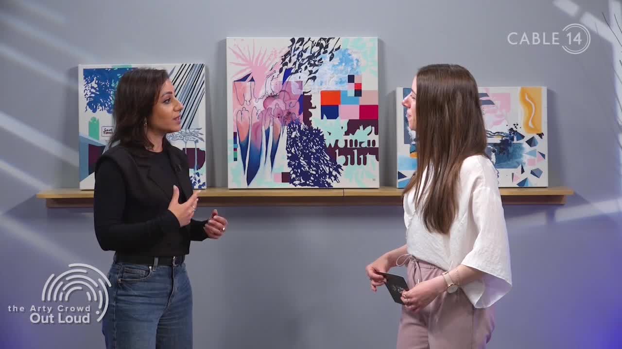 Two women stand in front of abstract paintings, one gesturing animatedly as she speaks. The Cable 14 logo is visible, suggesting this is a segment from Hamilton, Canada.
