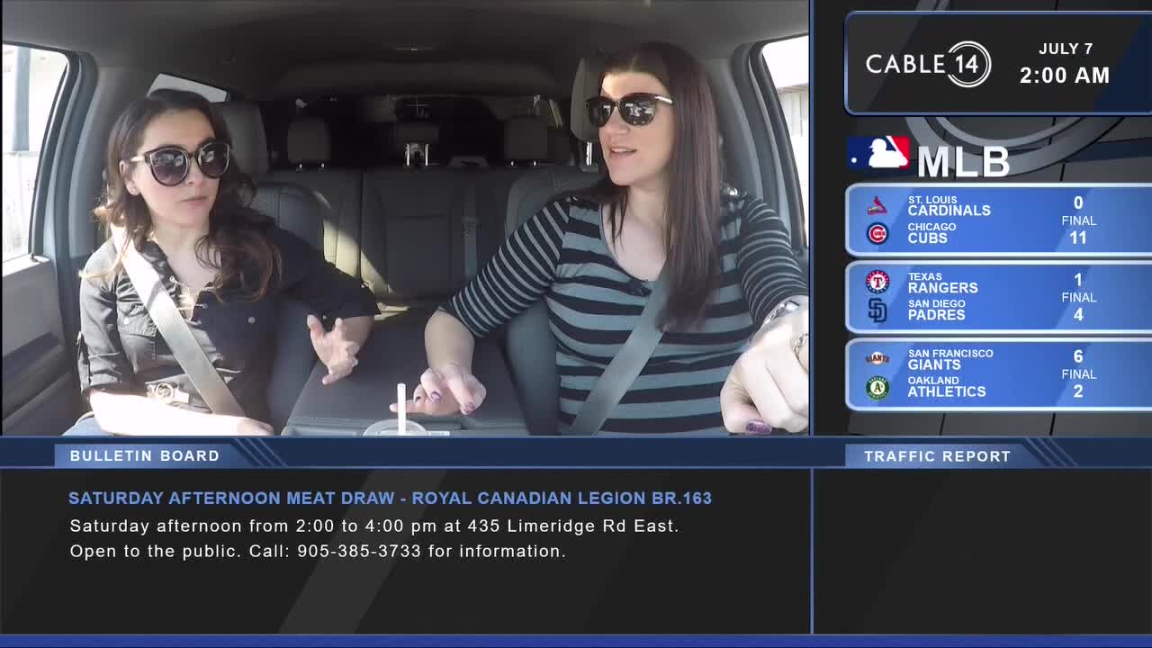 Two women in sunglasses are in a car, talking while a Cable 14 graphic displays baseball scores. The bulletin board below advertises a meat draw at the Royal Canadian Legion in Hamilton.
