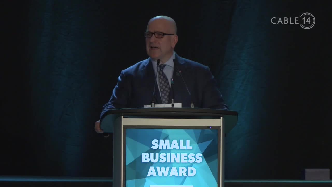 A man in a suit stands at a podium, speaking into a microphone with the Cable 14 logo visible in the corner. The podium displays the words "Small Business Award" on a screen.
