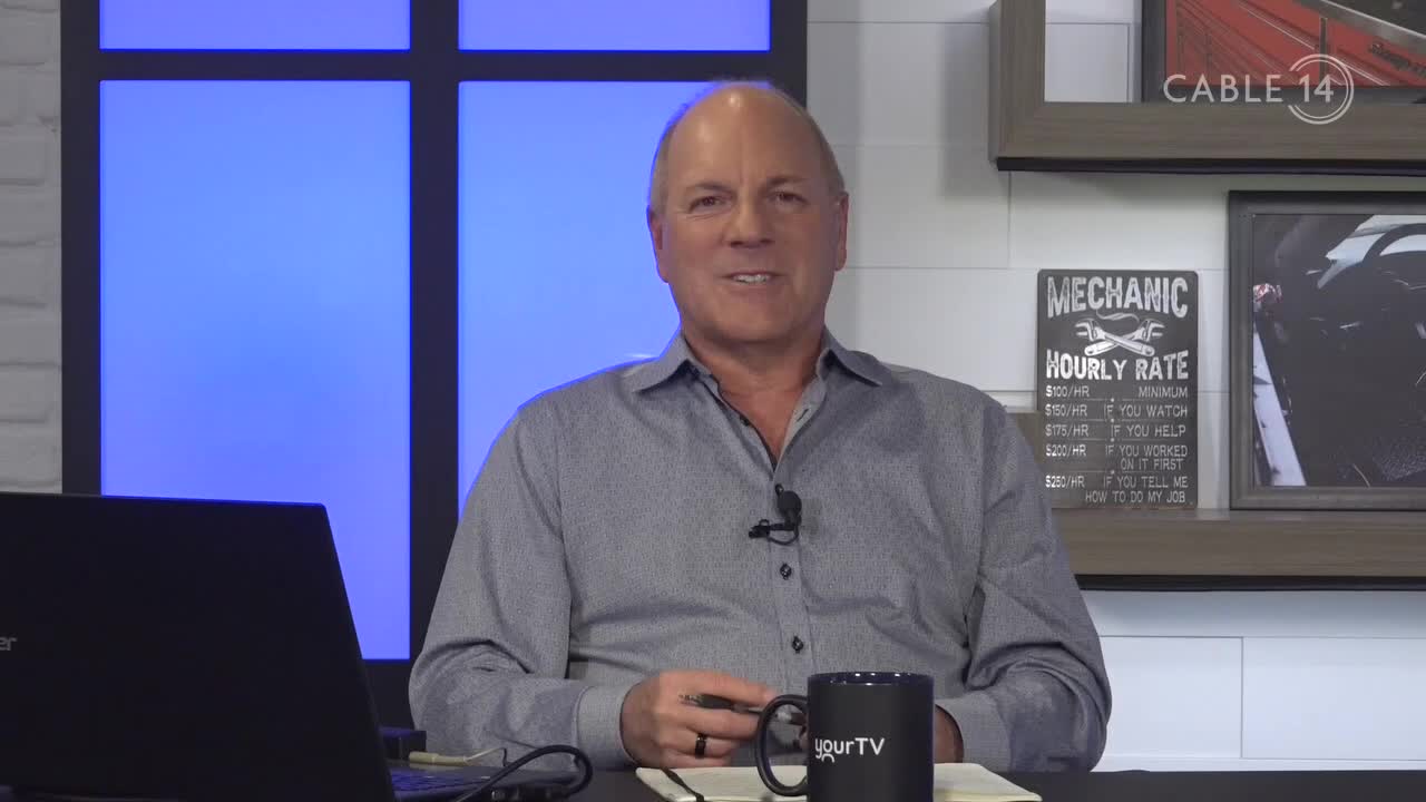 A man in a gray shirt sits at a desk, speaking into a microphone. The Cable 14 Hamilton logo is visible in the corner, and a mug with "yourTV" is on the desk.
