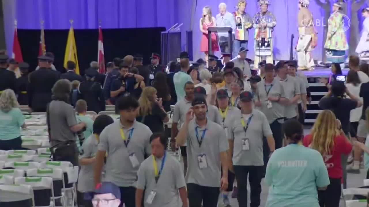 People in gray shirts with lanyards are walking toward the camera, while others mill about near a stage. The Cable 14 Hamilton logo is visible as the Canadian flag hangs in the background.
