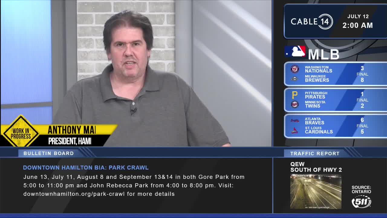 Anthony Mai, President of HAMI, is speaking on Cable 14, with baseball scores and a traffic report on the screen behind him. A "Work in Progress" sign is visible, and a local event in Downtown Hamilton is advertised.

