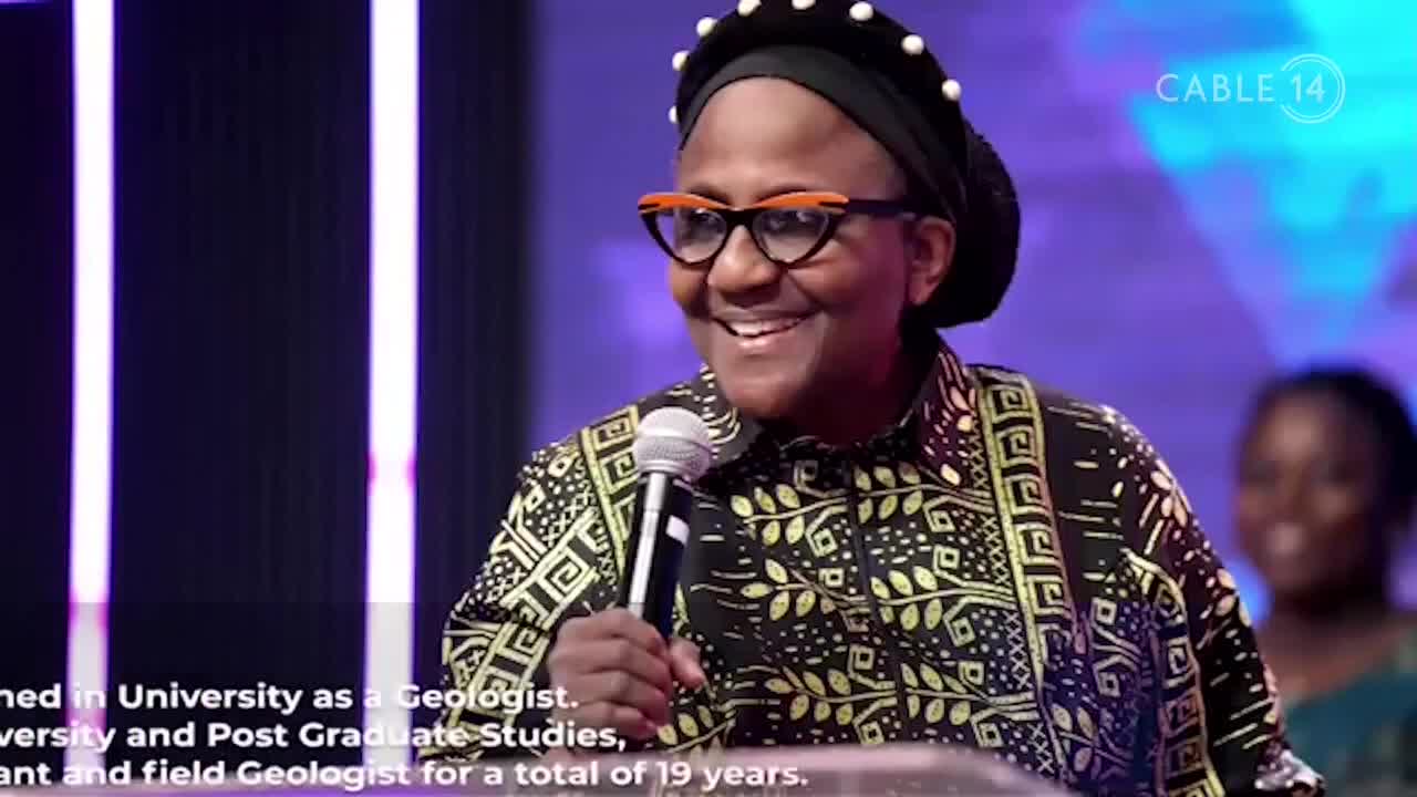 A woman in a patterned shirt and glasses smiles, holding a microphone on a Cable 14 Hamilton set. The background glows with vertical purple lights.
