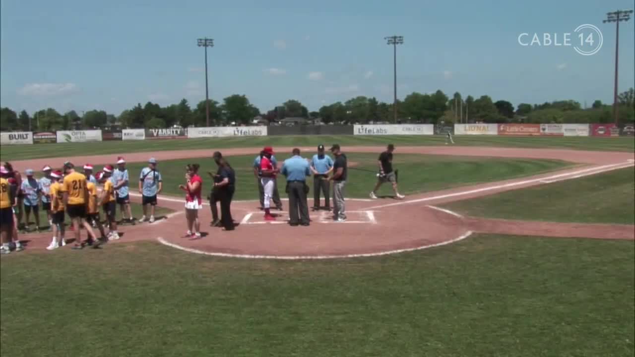 On a sunny day, a group of players in yellow jerseys huddle near the first base line, while officials and coaches gather around home plate. A Cable 14 logo is visible in the top right corner, suggesting this is a local broadcast from Hamilton, Canada.
