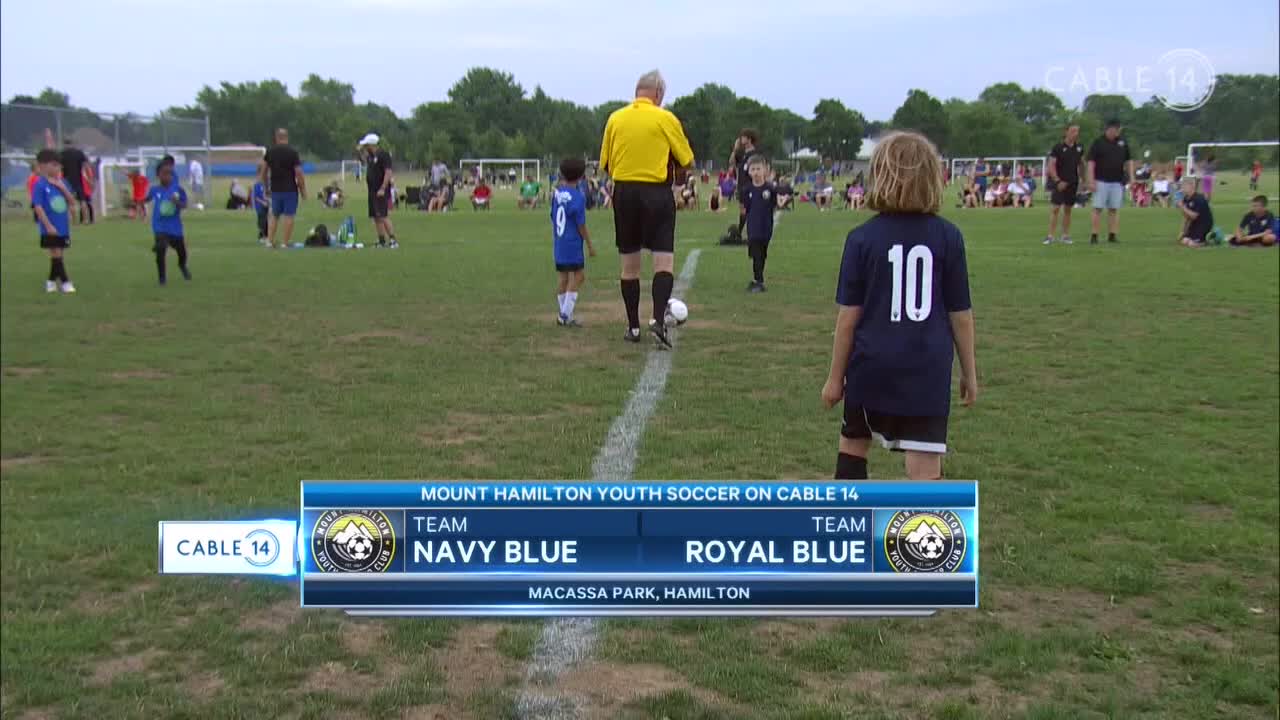 A referee in a yellow shirt stands over the ball, ready to start the game at Macassa Park. The Mount Hamilton Youth Soccer match between Navy Blue and Royal Blue is on Cable 14.
