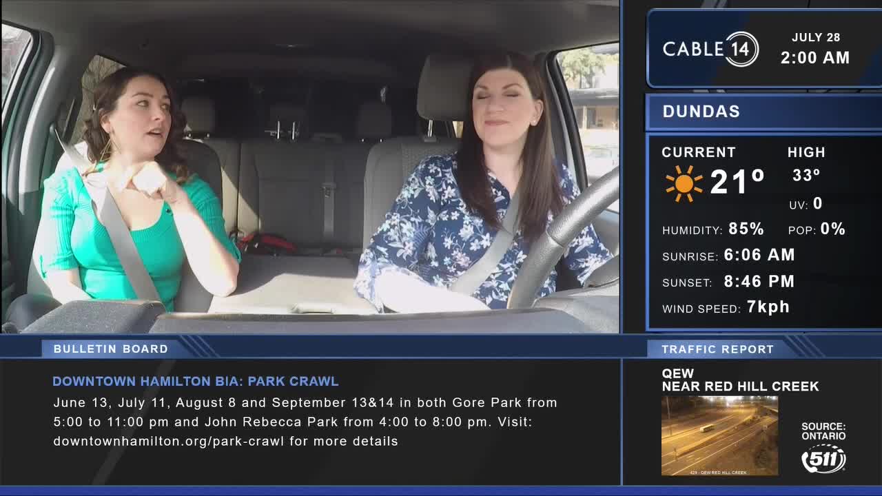 Two women are in a car, one driving and the other gesturing while talking. Cable 14 Hamilton is on the screen, with a weather report showing 21 degrees Celsius in Dundas.

