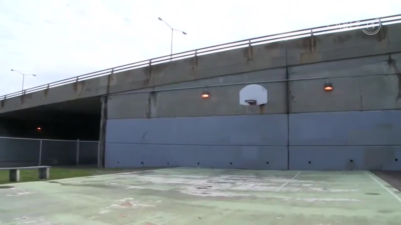 A basketball hoop is mounted on a concrete wall, beneath a bridge. The court is empty, and the sky is overcast.
