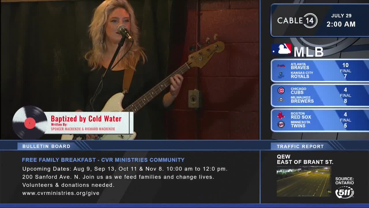 On Cable 14, a woman with blonde hair strums a white bass guitar, the "Baptized by Cold Water" title card displayed on the screen. The broadcast from Hamilton, Ontario, also shows MLB scores and a traffic report for the QEW.
