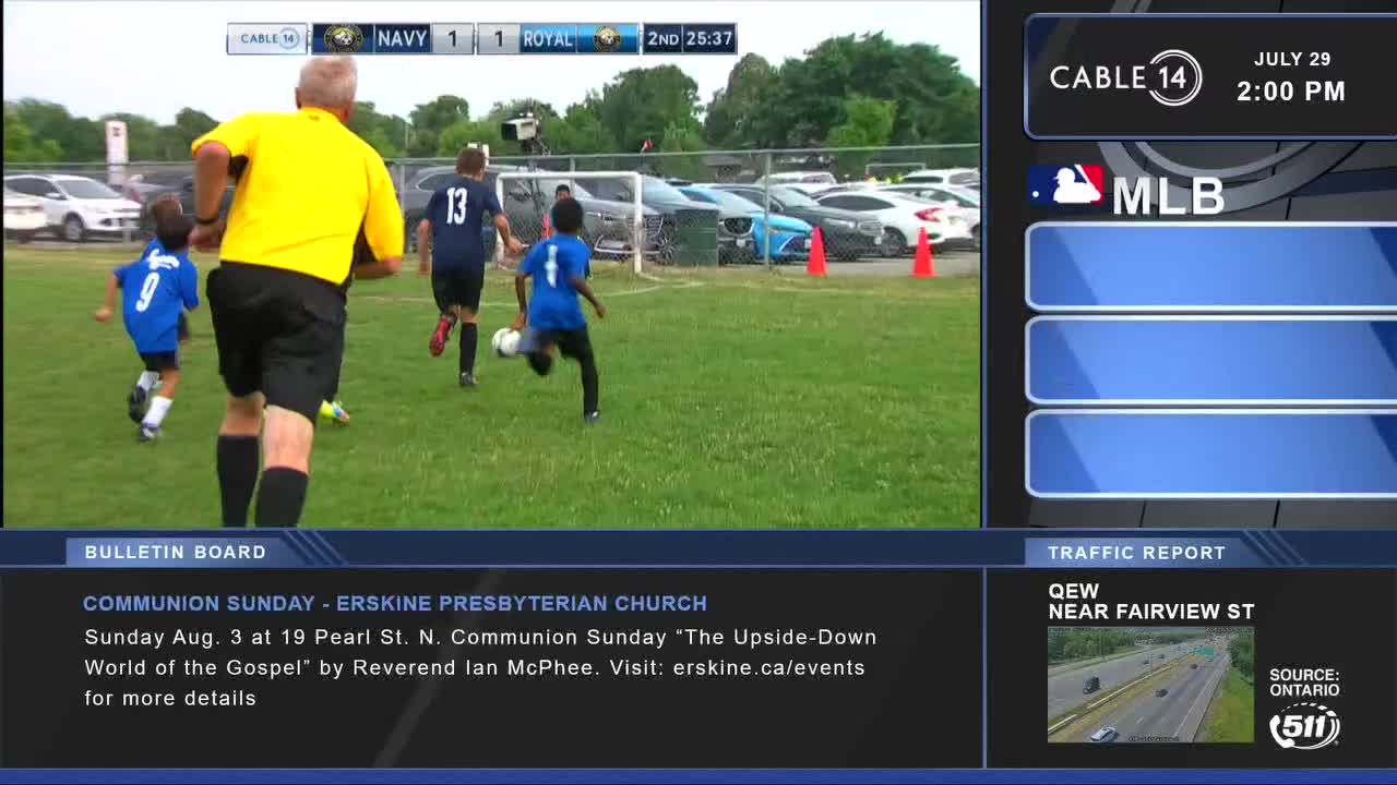 A player in blue sprints with the ball, chased by another in a dark jersey. The referee, in a bright yellow shirt, keeps pace as the game continues, as seen on Cable 14 Hamilton.

