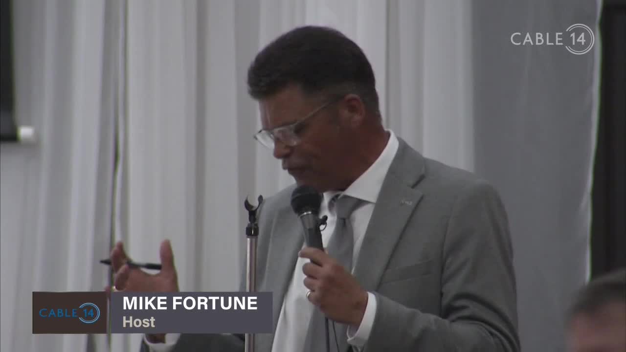 Mike Fortune, the Cable 14 host, is speaking into a microphone, gesturing with his left hand. He's wearing a grey suit and glasses, with the Cable 14 logo visible in the corner.
